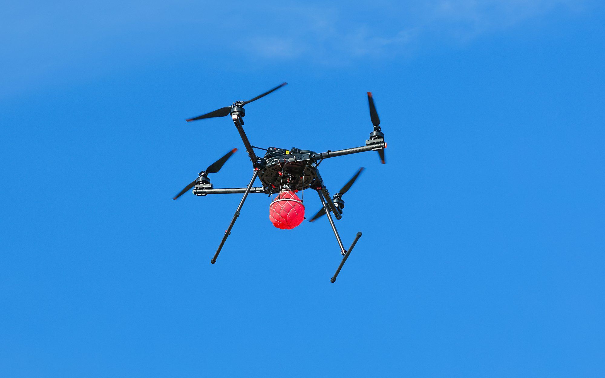 A black quadcopter drone flies with a red balloon beneath it.