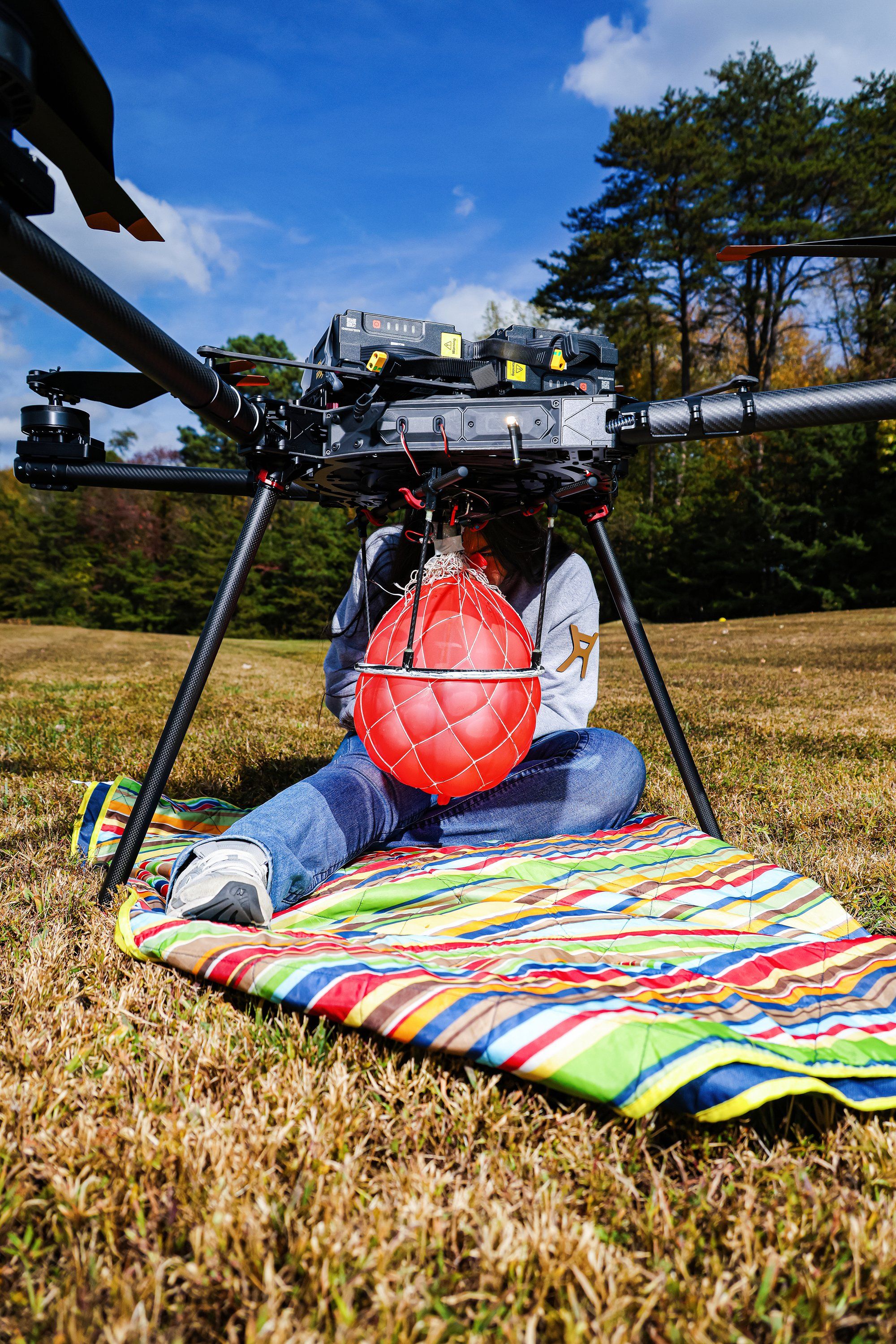 A woman sits behind a large drone. Her upper body is obscured by a red balloon attached to the droneu2019s underside.