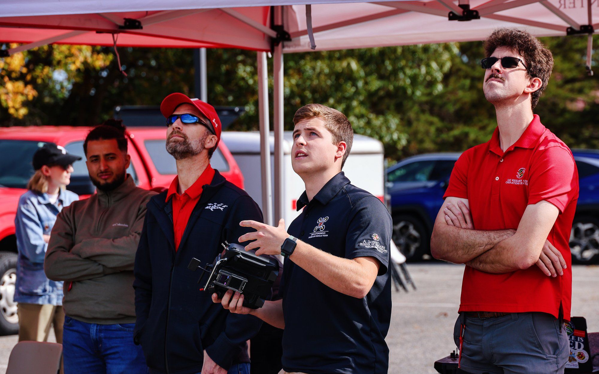 Four young men in the foreground and woman in the background stand in a parking lot. The right-most three men look skyward. One of them holds an electronic device.