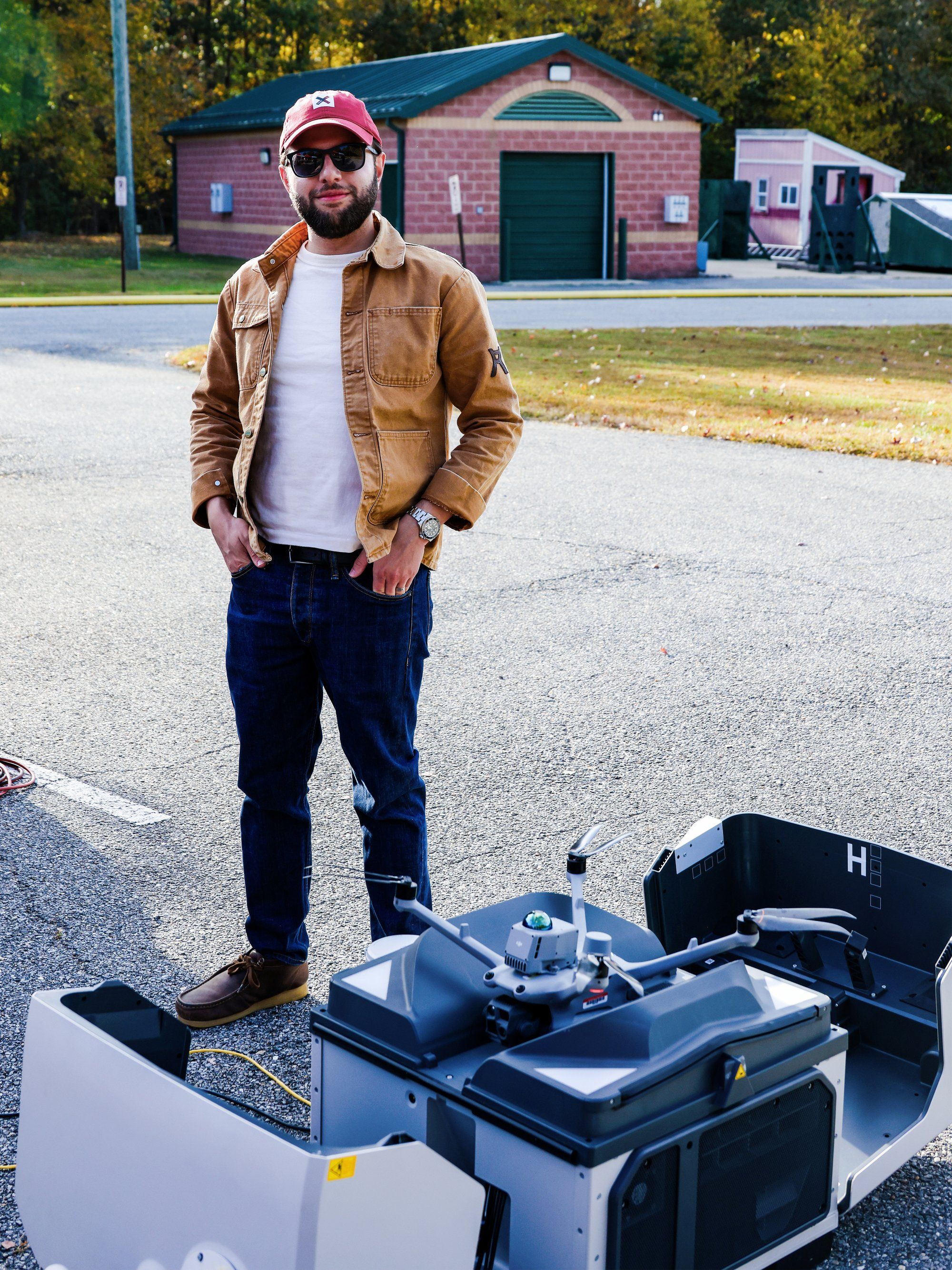 Person in a brown jacket and cap stands next to a large drone outdoors near a brick building.