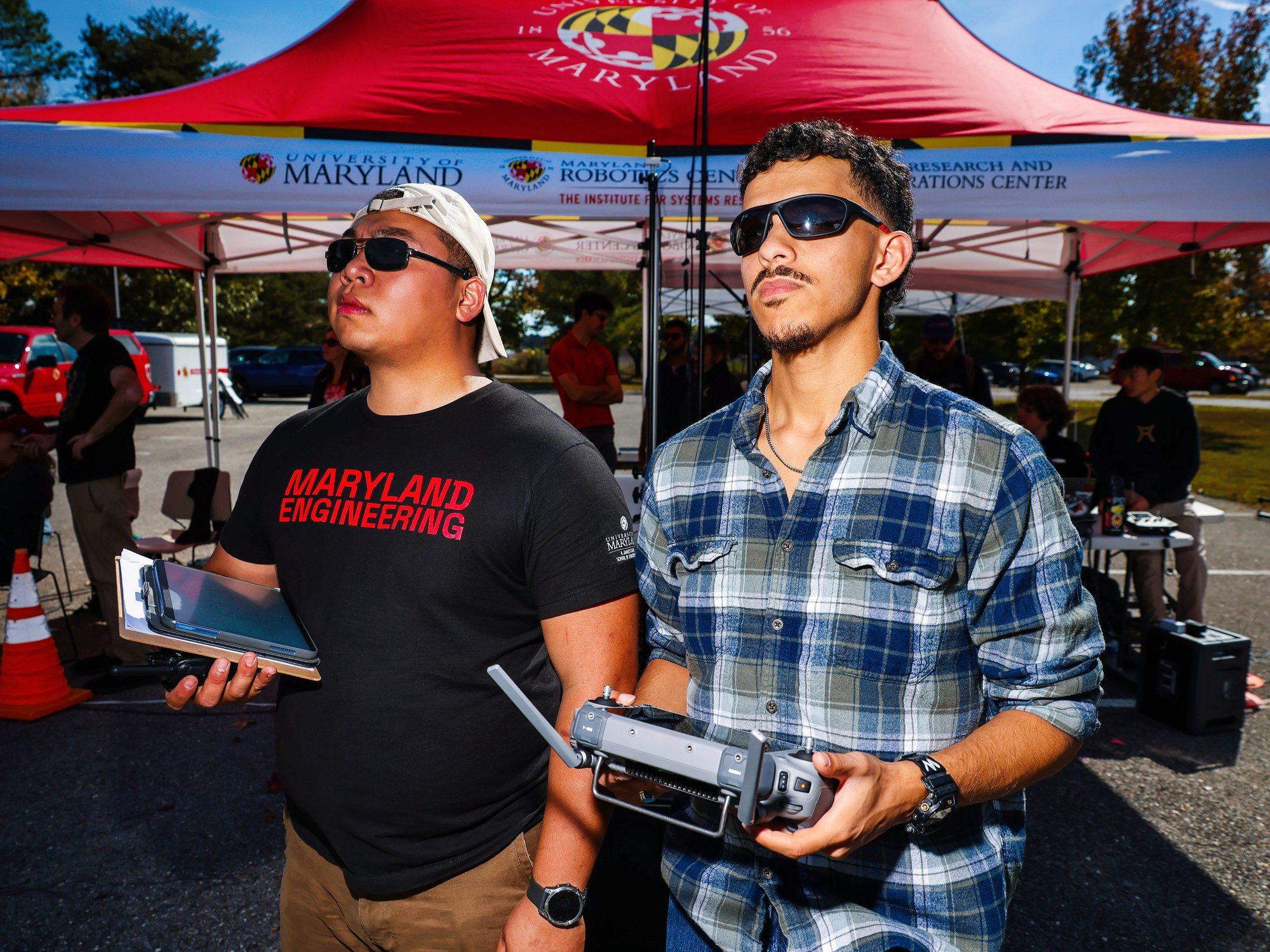 Two young men in sunglasses look up. One of the men holds a large remote control. 