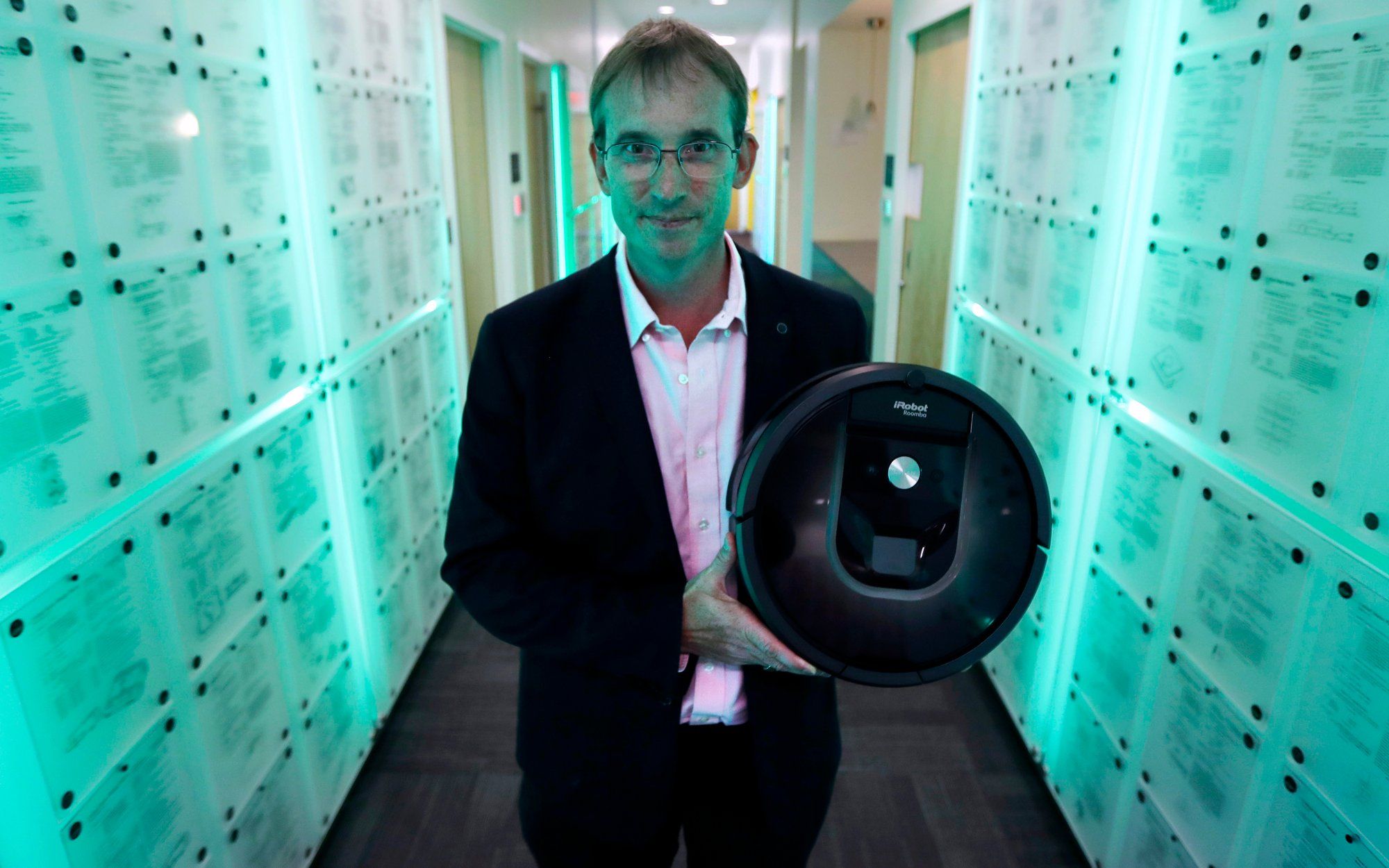 A suited man with short blonde hair and glasses holds a round black robot vacuum in a hallway covered in patents.